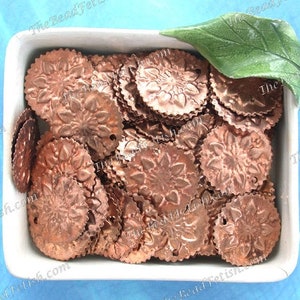 May include: A white ceramic dish filled with copper-colored metal charms. The charms are round and have a floral design. A green leaf is partially visible in the top right corner of the dish.