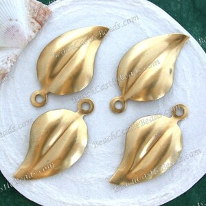 K&ouml;nnte beinhalten: Vier goldfarbene, metallene Anh&auml;nger in Blattform mit einem Loch oben zum Aufh&auml;ngen. Die Anh&auml;nger liegen auf einer wei&szlig;en Oberfl&auml;che.