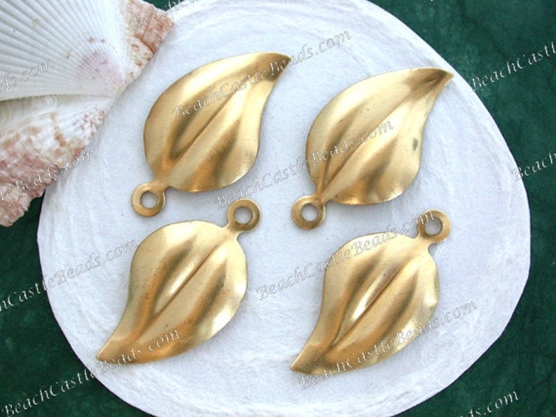 K&ouml;nnte beinhalten: Vier goldfarbene, metallene Anh&auml;nger in Blattform mit einem Loch oben zum Aufh&auml;ngen. Die Anh&auml;nger liegen auf einer wei&szlig;en Oberfl&auml;che.