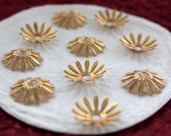 May include: A collection of gold-coloured metal bead caps, each shaped like a flower with petal-like edges and a central hole. They are displayed on a white, textured surface, with a blurred burgundy background.
