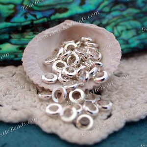 May include: A collection of small, shiny silver-colored metal ring beads. The beads are scattered inside a white seashell, resting on a white crocheted surface. The background features a blurred, iridescent green and blue surface.