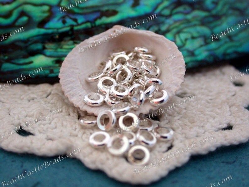 May include: A collection of small, shiny silver-colored metal ring beads. The beads are scattered inside a white seashell, resting on a white crocheted surface. The background features a blurred, iridescent green and blue surface.