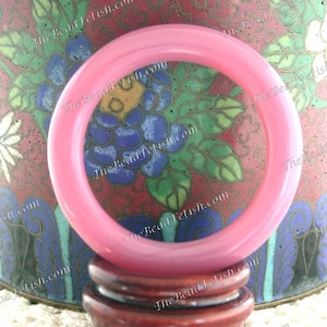May include: A translucent pink bangle bracelet. The bracelet is circular and sits on a wooden stand. The background features a decorative vase with floral patterns in blue, green, and red.