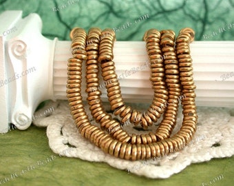 May include: Strands of golden-colored, round beads are arranged in a layered pattern. The beads have a metallic finish and are strung together. The beads are displayed on a white, decorative surface with a green background.