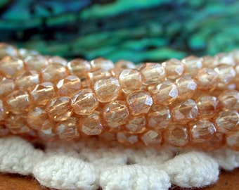 May include: A strand of iridescent peach-colored glass beads. The beads are faceted and have a shimmery finish. The beads are strung together on a white crocheted surface.