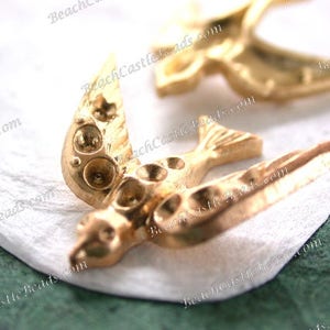 May include: Two gold-tone metal bird charms with detailed wings and bodies. The charms are on a white surface.
