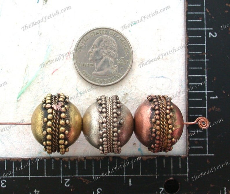 May include: Three round beads with a metallic finish. The beads are made of metal and have a textured surface. The beads are gold, silver, and copper in color. The beads are approximately 1 inch in diameter.