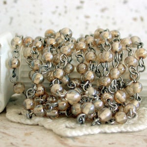 May include: A strand of gold-toned glass beads strung on a silver chain. The beads are round and have a slight iridescent sheen.