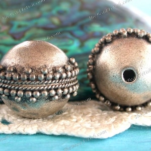 May include: Two silver metal beads with a textured surface and a beaded trim. The beads have a hole in the center for stringing.