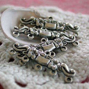 May include: Close-up of four silver-toned metal clasps with floral and scroll designs. Each clasp features a central flower, curved accents, and multiple small loops for attachment. The clasps are arranged in a white seashell on a white crocheted surface.