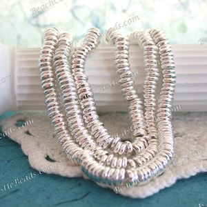 May include: Several strands of silver-colored, ring-shaped beads are arranged on a white, crocheted doily. The beads have a shiny, metallic finish and are tightly strung together. The background includes a white architectural element and a turquoise surface.