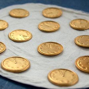 May include: A white surface with 11 gold-coloured clock face charms. The charms are round and have black numbers and hands.