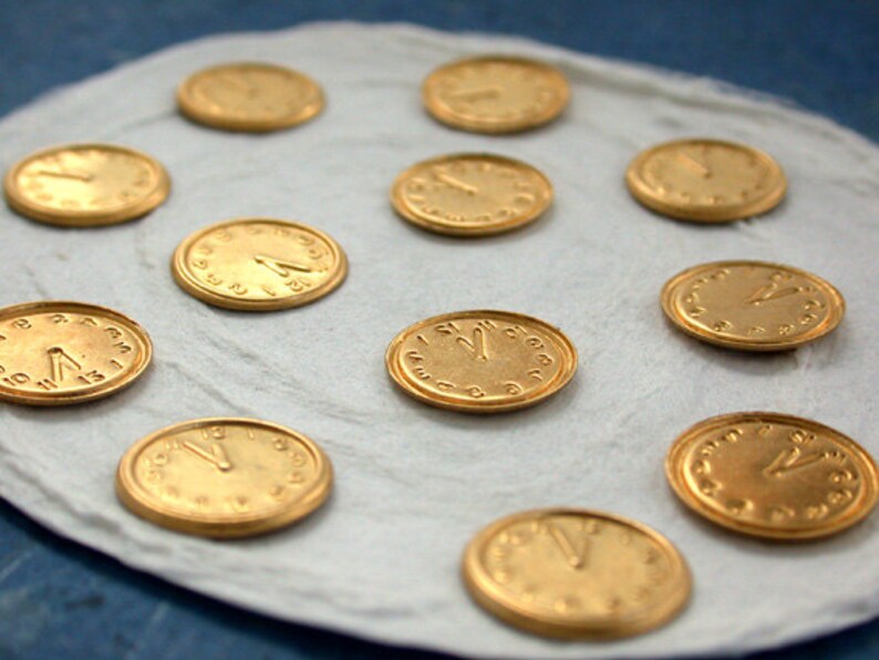 May include: A white surface with 11 gold-coloured clock face charms. The charms are round and have black numbers and hands.
