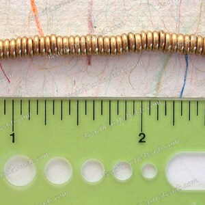 Op de afbeelding: Close-up van een streng goudkleurige metalen kralen. De kralen zijn cilindrisch en aan elkaar geregen. Een liniaal eronder toont de afmetingen in centimeters.
