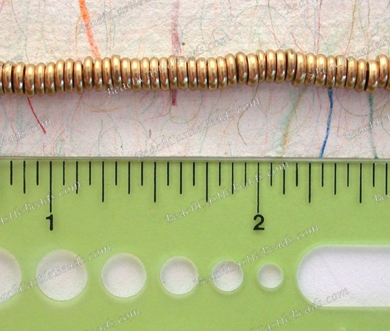 Op de afbeelding: Close-up van een streng goudkleurige metalen kralen. De kralen zijn cilindrisch en aan elkaar geregen. Een liniaal eronder toont de afmetingen in centimeters.