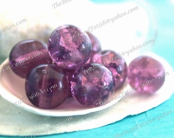 May include: A selection of translucent, round, purple beads with internal veining, presented in a white, shell-shaped dish. The beads have a glossy finish and varying shades of purple. The background is a light blue.