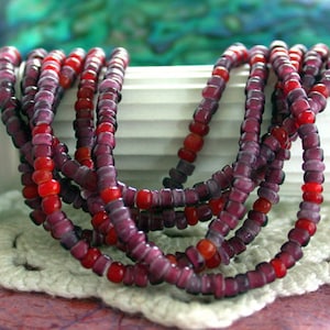 May include: Strands of beaded necklaces in shades of red and purple. The beads are small and round, strung together to create multiple layers. The necklaces are draped over a white surface with a crocheted edge, creating a layered effect.