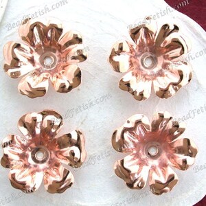 May include: Four rose-gold colored metal flower embellishments with a hole in the center. The flowers are on a white surface.