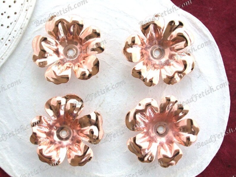 May include: Four rose-gold colored metal flower embellishments with a hole in the center. The flowers are on a white surface.