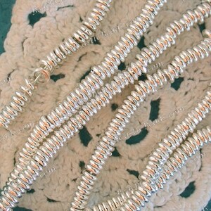 May include: Close-up of several strands of shiny, silver-colored beads. The beads have a ridged, spiral design and are strung together. The background is a cream-colored, crocheted doily.