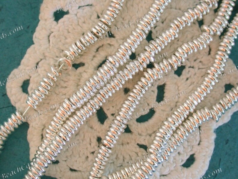 May include: Close-up of several strands of shiny, silver-colored beads. The beads have a ridged, spiral design and are strung together. The background is a cream-colored, crocheted doily.