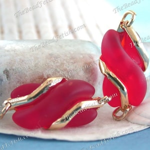 May include: Red glass pendant earrings with gold-colored metal accents. The earrings have a unique, flowing design. The background includes a seashell and a blurred blue backdrop.