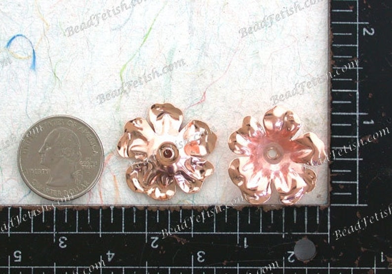 May include: Two rose gold metal flower embellishments, each with five petals and a hole in the center. The flowers are approximately 1 inch in diameter and are shown next to a ruler.