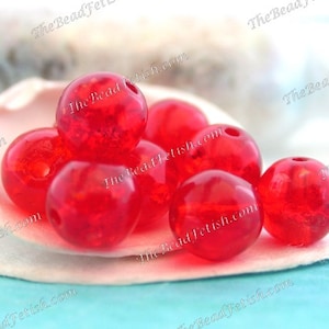 May include: A collection of translucent red beads with a hole through the center, resting in a white seashell. The beads have a slightly textured appearance and are round in shape. The background is a soft blue.