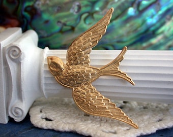 Puede incluir: Un broche de metal dorado en forma de pájaro con patrones de plumas detallados. El pájaro está en vuelo con las alas extendidas.