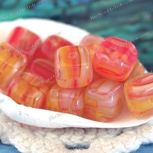 May include: A collection of translucent, square-shaped beads in shades of orange, yellow, and red, displayed in a white seashell. The beads have a striped pattern and a glossy finish, perfect for jewelry making.