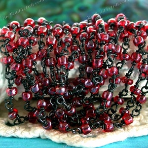 May include: A close-up of a red glass bead chain with black metal chain links. The chain is draped over a white surface.