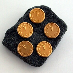May include: Five gold-coloured clock face pins with black hands and black numbers on a dark grey rock.