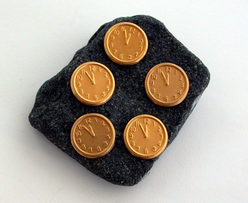May include: Five gold-coloured clock face pins with black hands and black numbers on a dark grey rock.