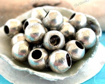 May include: A close-up of a pile of silver metal beads with a hole in the centre of each bead. The beads are in a white bowl with a scalloped edge.