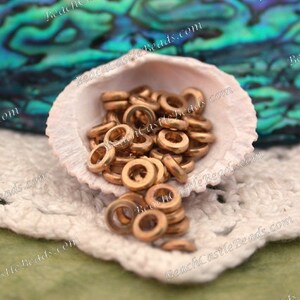 Op de afbeelding: Een verzameling kleine, bronskleurige metalen ringkralen wordt getoond in een witte zeeschelp. De kralen zijn verspreid, sommige vallen eruit. De achtergrond omvat een groen-blauw iriserend oppervlak en een wit gehaakt kleedje.