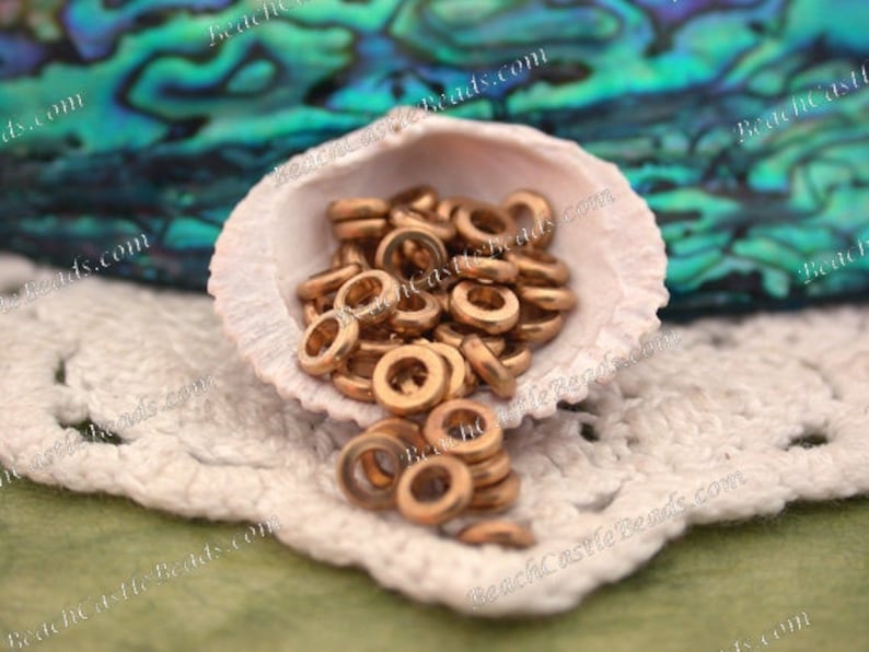 Op de afbeelding: Een verzameling kleine, bronskleurige metalen ringkralen wordt getoond in een witte zeeschelp. De kralen zijn verspreid, sommige vallen eruit. De achtergrond omvat een groen-blauw iriserend oppervlak en een wit gehaakt kleedje.
