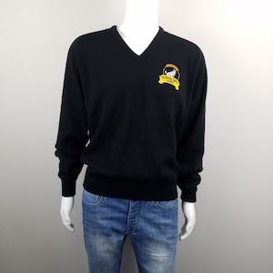 May include: A navy blue long-sleeve V-neck sweater featuring an embroidered patch on the left chest. The patch displays a white wolf graphic above a yellow banner with the text "NORTH WOLF LIGHT" in black lettering. The sweater is paired with blue jeans.