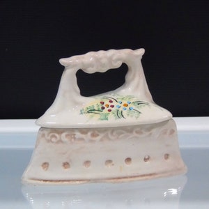 May include: A ceramic iron-shaped trinket box in a cream color. The lid features a floral design with red, blue, and yellow accents. The base has a row of small holes and a decorative pattern. The handle has a curved design.