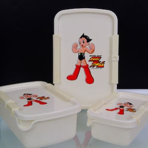 May include: A set of three off-white plastic food containers. Each container features a cartoon image of Astro Boy in red boots and a black outfit. The largest container is rectangular, and the other two are square-shaped.
