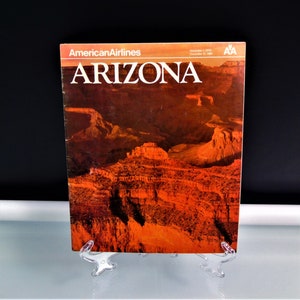 May include: A vintage American Airlines magazine cover featuring the word "ARIZONA" in large white letters. The cover displays a vibrant image of the Arizona landscape, showcasing red rock formations under a warm, orange sky. The magazine is displayed on a clear plastic stand.