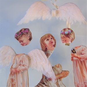 May include: A collage of angel-themed paper cutouts. The image shows detached angel wings made of white feathers, and several cherubic faces adorned with floral crowns. The angels are depicted in flowing white gowns, some holding objects.