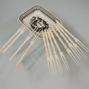 May include: A set of ten off-white, two-pronged cocktail forks with textured, beaded handles, arranged in a fan shape inside a small, rectangular, cream-colored ceramic dish featuring a black and white landscape illustration on its interior base.