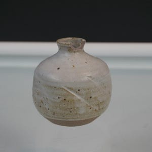 May include: A small, round ceramic vase with a narrow neck and a small notch on the rim. The body features a mottled off-white glaze with visible brown speckles and subtle diagonal indentations, transitioning to a plain, unglazed base.