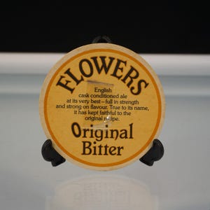 Puede incluir: Un posavasos redondo Flowers Original Bitter de color beige con texto negro. El posavasos tiene las palabras "FLOWERS" en la parte superior, con texto debajo que describe las cualidades de la cerveza. Las palabras "Original Bitter" est&aacute;n en la parte inferior.