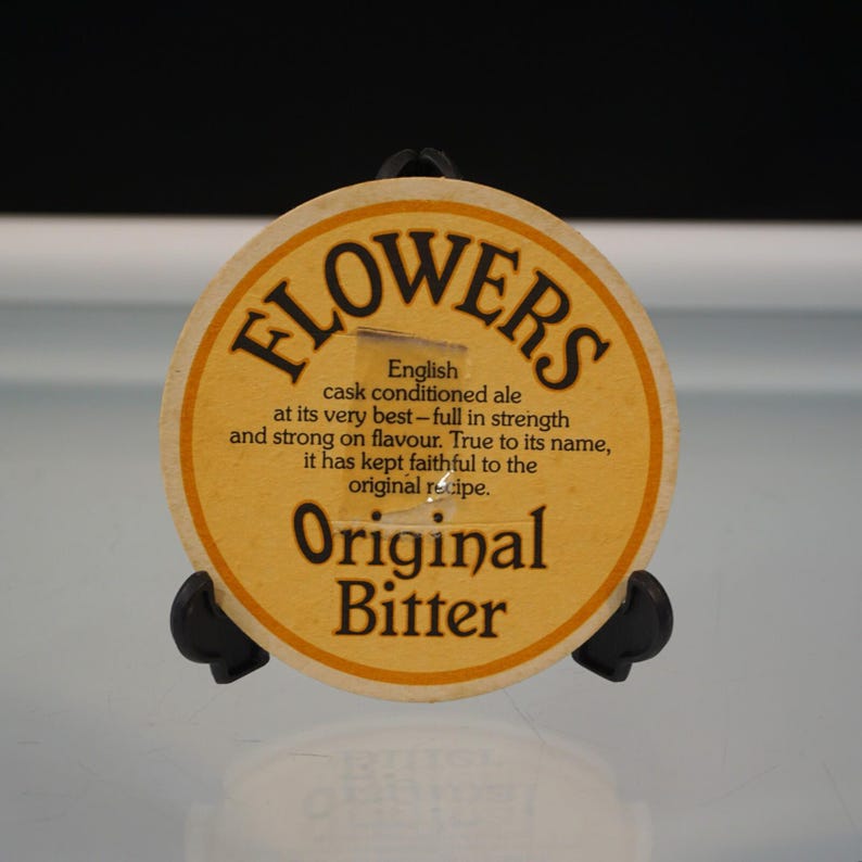 Puede incluir: Un posavasos redondo Flowers Original Bitter de color beige con texto negro. El posavasos tiene las palabras "FLOWERS" en la parte superior, con texto debajo que describe las cualidades de la cerveza. Las palabras "Original Bitter" est&aacute;n en la parte inferior.