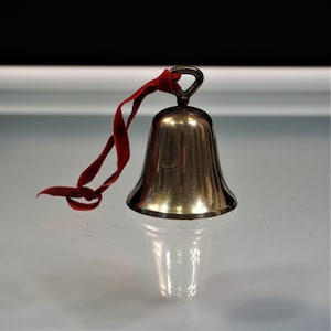 May include: A polished metallic bell with a smooth, flared body and a small loop at the top. A thin, vibrant red velvet ribbon is tied through the loop, hanging loosely to the side. The bell has a reflective surface that catches light.