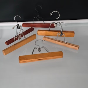 May include: A collection of five wooden pant hangers featuring horizontal bars with metal clamping mechanisms and swivel hooks. The hangers vary in wood tones from light natural to dark reddish-brown, each equipped with silver-toned metal hardware.