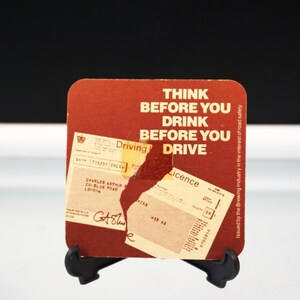 Puede incluir: Un posavasos cuadrado, de color marr&oacute;n rojizo, con el texto "THINK BEFORE YOU DRINK BEFORE YOU DRIVE" en blanco. El posavasos tiene una secci&oacute;n rasgada que revela una licencia de conducir y otros documentos. El posavasos est&aacute; sobre un soporte negro.