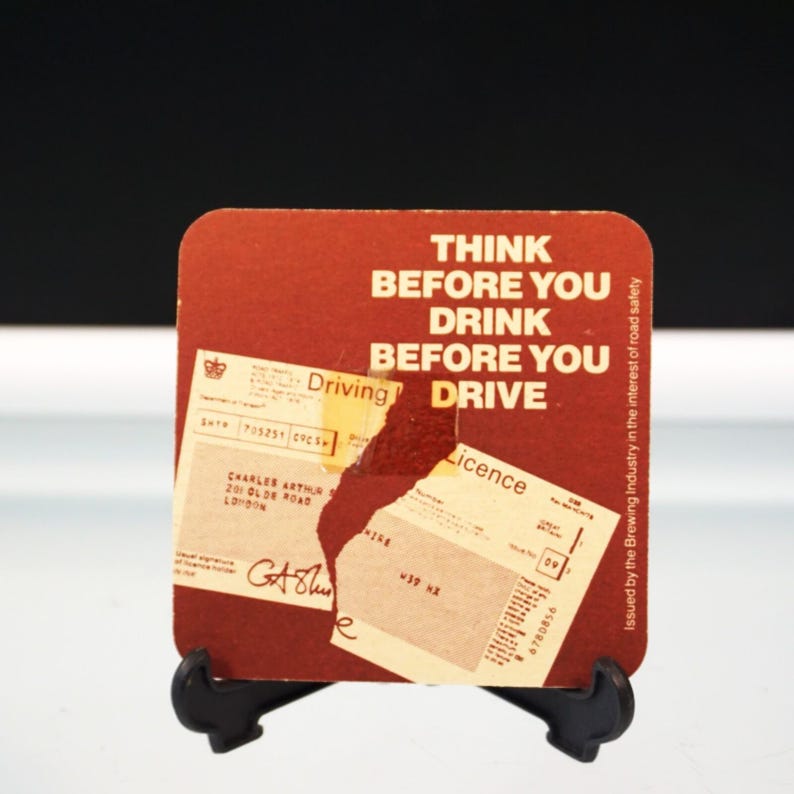 Puede incluir: Un posavasos cuadrado, de color marr&oacute;n rojizo, con el texto "THINK BEFORE YOU DRINK BEFORE YOU DRIVE" en blanco. El posavasos tiene una secci&oacute;n rasgada que revela una licencia de conducir y otros documentos. El posavasos est&aacute; sobre un soporte negro.