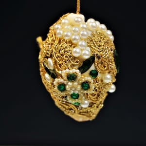 May include: An egg-shaped ornament with a gold filigree design, decorated with faux pearls and emerald-green gemstones. The ornament hangs from a gold string against a black backdrop, highlighting its intricate details.